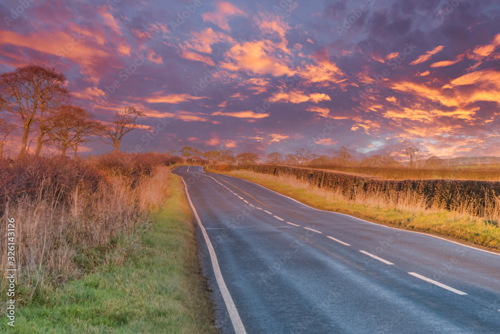 Scottish Country Road in Autumn as the sun goes down leaving a dramamtic blazing red sky. The far distance is Hazy due to the Reflected Light and mist.