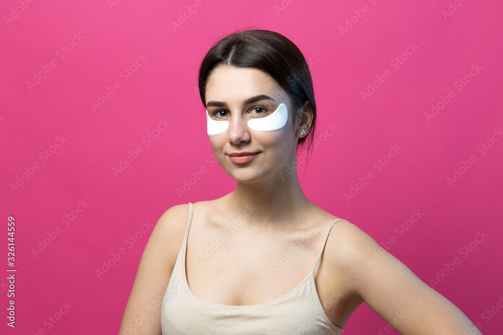 Close up portrait of pretty attractive girl with naked shoulders using patches under eyes. Standing over pink background. Close up portrait of pretty attractive girl with naked shoulders using patches under eyes. Standing over pink background.