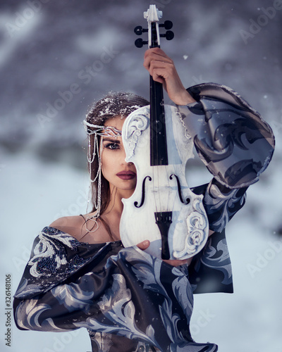 Portrait amazing cute slim young woman in chic fairy tale image in gray puffy dress and with crown on her head holds beautiful white violin and bow against background of white snow covered forest