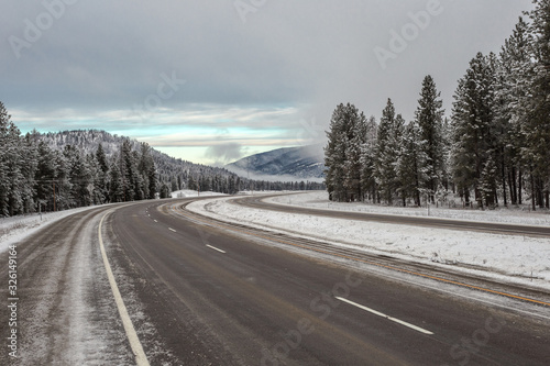 Empty curving highway surrounded by snow and tall evergreen trees leading to fog shrouded mountain