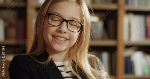 Close up of blonde Caucasian pretty teen small girl in glasses finishing to read book and closing it. Beautiful little blond schoolgirl smiling to camera in library. Portrait of cute smiled kid.