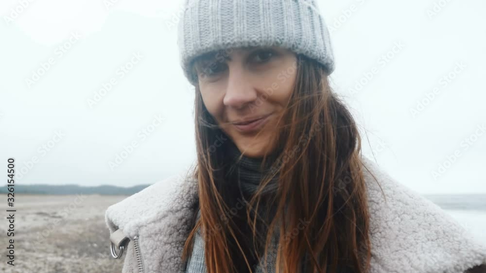 Close-up portrait of beautiful happy female tourist smiling at camera, posing at cold windy winter sea beach slow motion Close-up portrait of beautiful happy female tourist smiling at camera, posing at cold windy winter sea beach slow motion