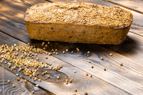Gluten-free buckwheat bread with a golden brown crust, sprinkled with sesame seeds, lies on a wooden table. Healthy homemade recipe. Grains of green buckwheat are scattered nearby. Copy space