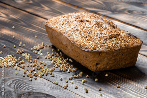 Gluten-free buckwheat bread with a golden brown crust, sprinkled with sesame seeds, lies on a wooden table. Healthy homemade recipe. Grains of green buckwheat are scattered nearby. Copy space