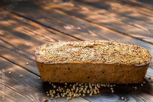 Gluten-free buckwheat bread with a golden brown crust, sprinkled with sesame seeds, lies on a wooden table. Healthy homemade recipe. Grains of green buckwheat are scattered nearby. Copy space