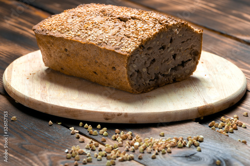 A loaf of freshly baked buckwheat bread lies on a round kitchen board on a brown wooden table. A handful of green buckwheat seeds are scattered nearby. Harmless baking for vegans and vegetarians