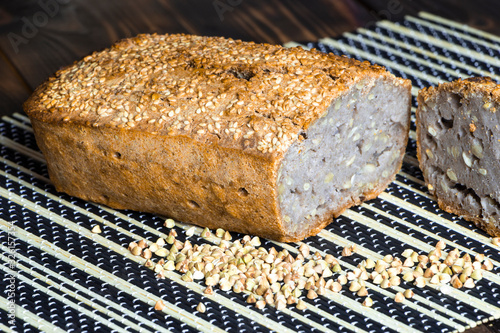 Vegan buckwheat bread. Freshly baked traditional bread on a bamboo serving mat on wooden table. Scattered grains and a slice near it. Selective focus. Healthy eating. Homemade natural food