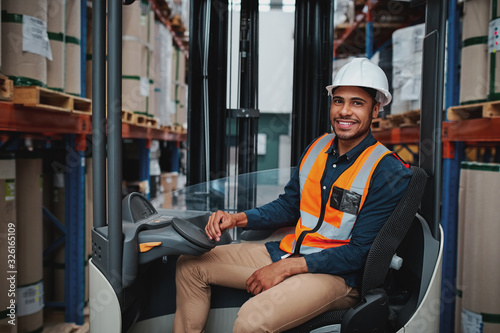 Happy young african forklift manager transporting goods from one shelf to another while looking in camera in warehouse