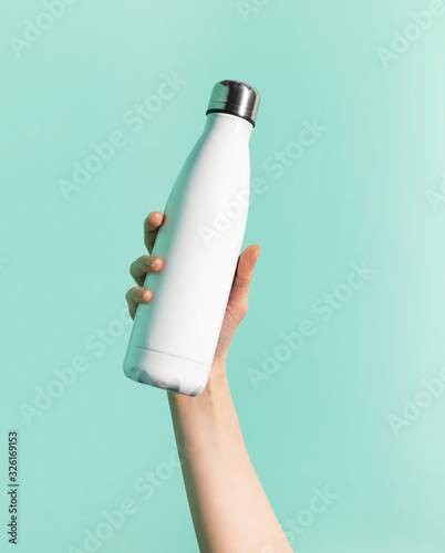 Close-up of female hand holding white reusable steel stainless thermo water bottle isolated on background of cyan, aqua menthe color. Plastic free. photo