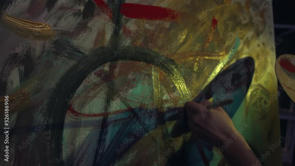 A young female artist is creating an abstract painting. She is working in the loft space with dimmed lights. She is using oil paint and a brush.