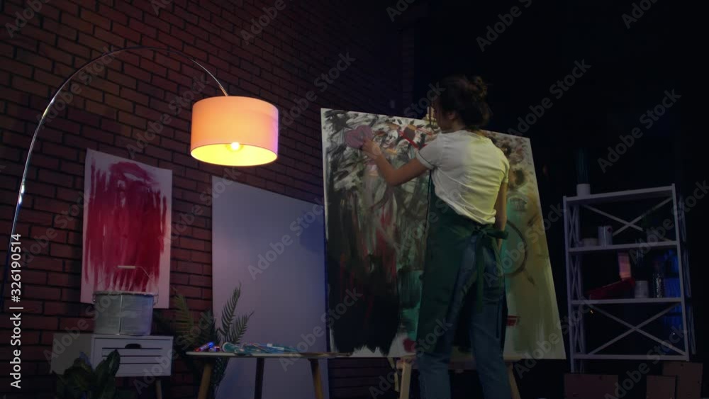 A young female artist is creating an abstract painting. She is working in the loft space with dimmed lights. She is using oil paint and a brush.