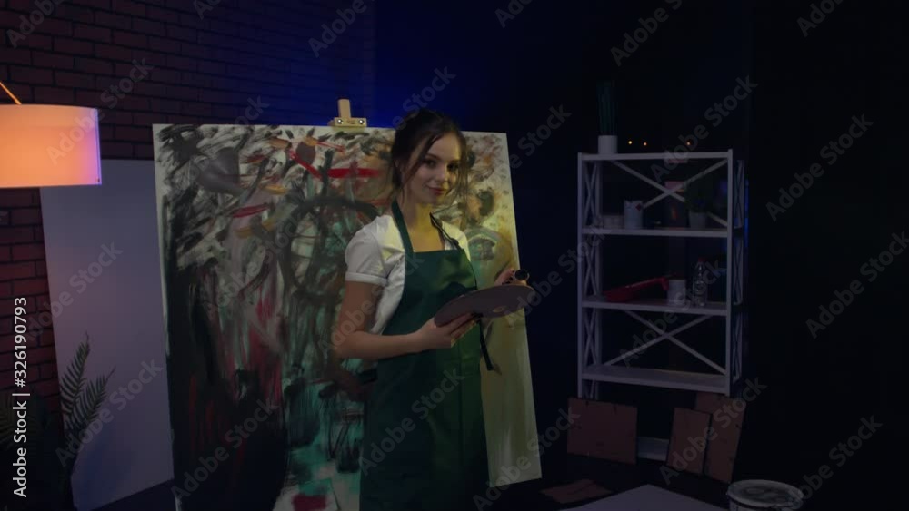 A young female artist is creating an abstract painting. She is working in the loft space with dimmed lights. She is using oil paint and a brush.