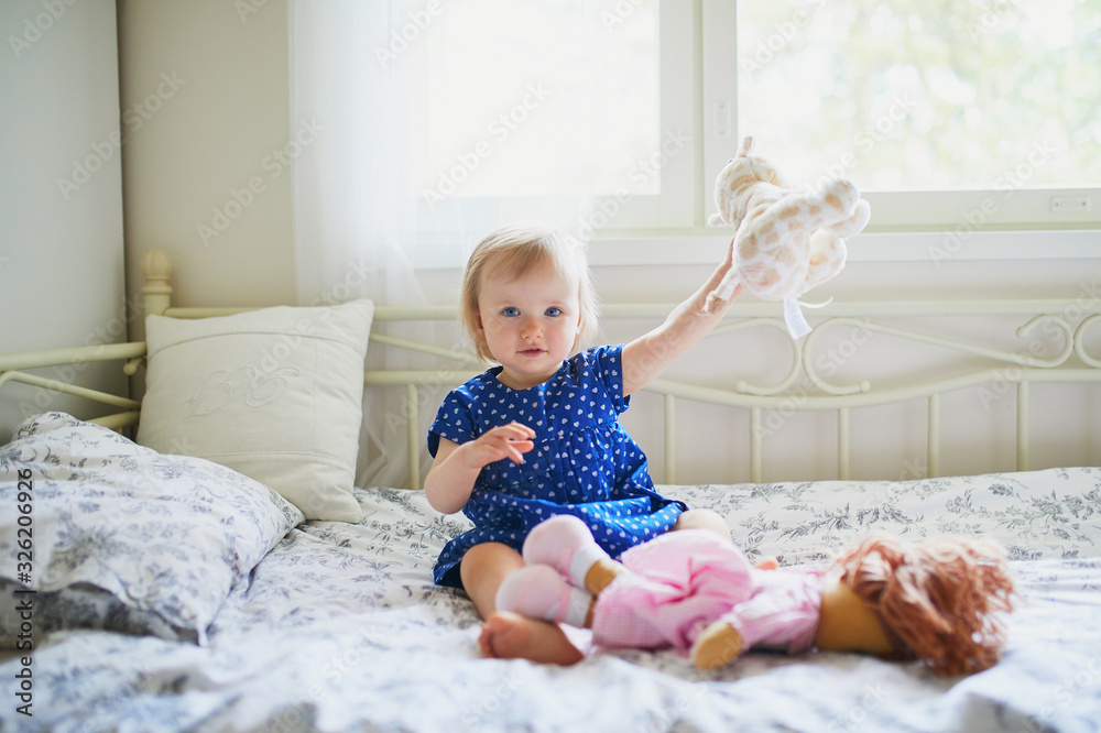Adorable baby girl in blue dress sitting on bed and playing with doll, teddy bear and dog Adorable baby girl in blue dress sitting on bed and playing with doll, teddy bear and dog