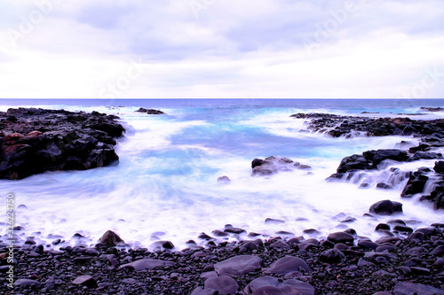 Ocean bay with rocky shores and azure blue water washing dark stones. Aerated foaming sea water in long exposure blue hour artistic vision of the wild sea.