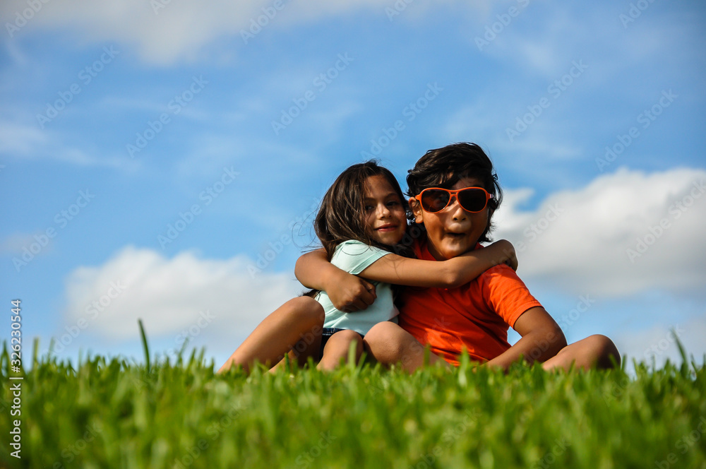 cuban indian bro sis joking pucker lips hugging spring orange blue shirt sitting on grass blue clouds tanned skin silly kids fun cuban indian bro sis joking pucker lips hugging spring orange blue shirt sitting on grass blue clouds tanned skin silly kids fun