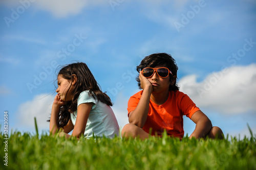 cuban indian brother sister boy girl sitting on grass angry bored hand to chin frustrated back to back blue sky white clouds orange shirt blue shirt hair blowing thinking