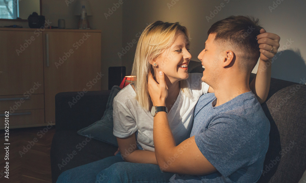 Cheerful caucasian boy embracing his blonde girlfriend while sitting on the sofa and smiling beautifully Cheerful caucasian boy embracing his blonde girlfriend while sitting on the sofa and smiling beautifully