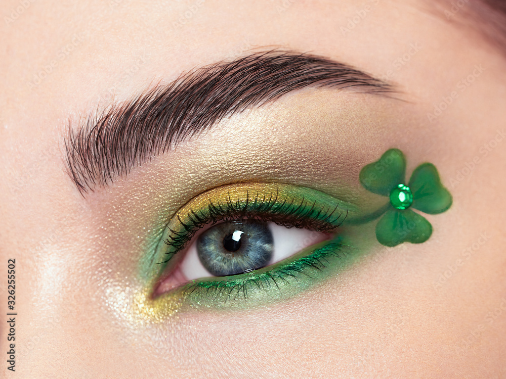 Conceptual photo of St. Patrick's Day. The eye of the young beautiful woman with bright green shadows and expressive eyebrows. Shamrock patterns. Holiday makeup - obrazy, fototapety, plakaty 