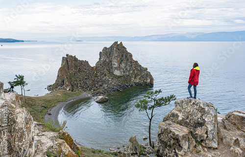 A girl in a red jacket stands on a rock on the shore of lake Baikal