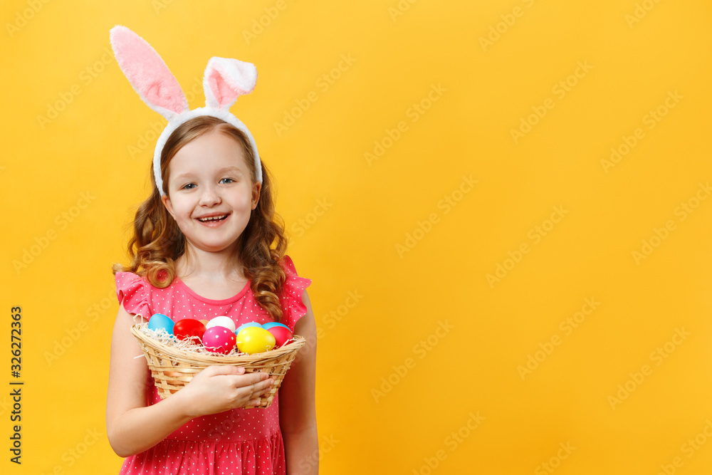 Happy beautiful cute little girl in easter bunny ears holds a basket with eggs. Spring close-up portrait of a child on a yellow background. Copy space