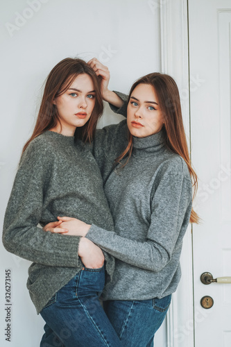 Young beautiful long hair girls sisters twins in casual grey sweaters and jeans on background of the white door