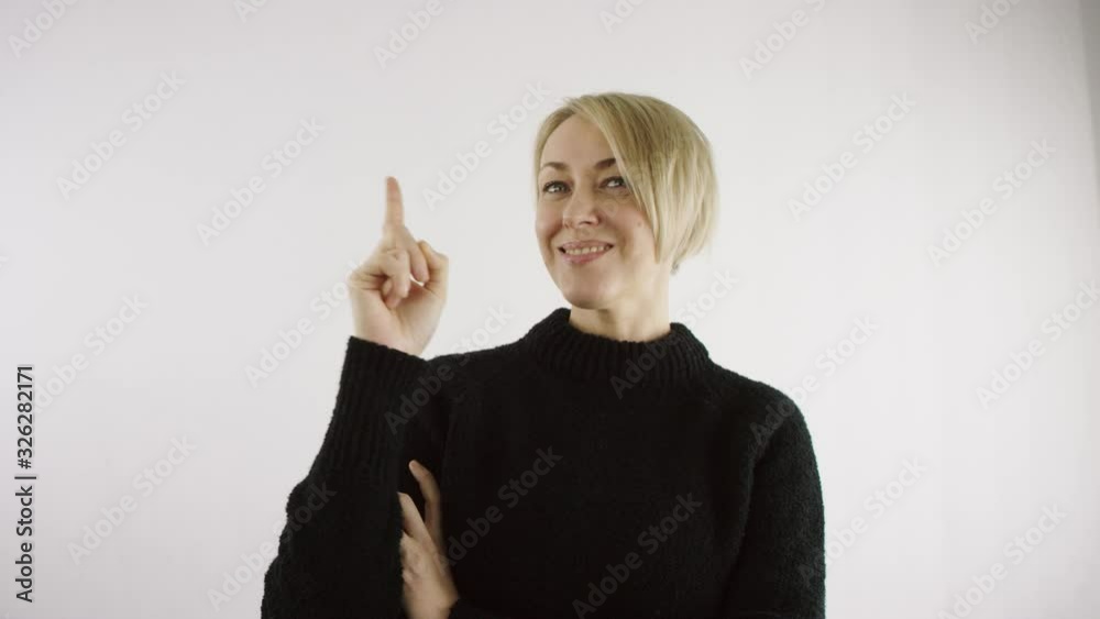 Adult middle aged caucasian blond short haired woman is points her index finger up to make a note on white background in bright interior Adult middle aged caucasian blond short haired woman is points her index finger up to make a note on white background in bright interior
