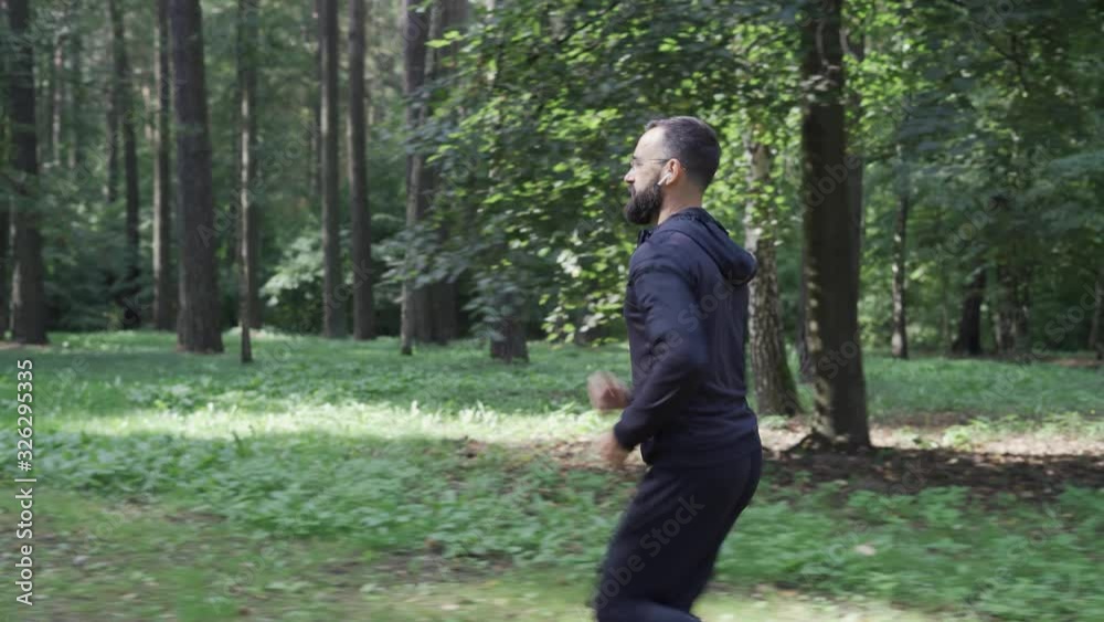 Adult man running through summer forest, camera following, runner in wireless headphones, healthy lifestyle.