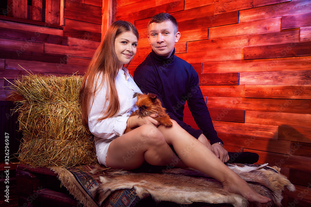 Beautiful girl in white shirt and guy in black sweater with small dog on the couch with hay in the barn or in the hayloft with wooden walls. Couple in rustic interior during photo shoot