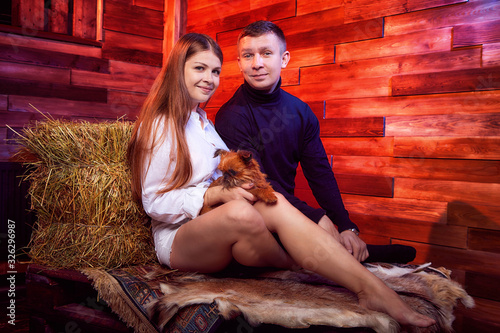 Beautiful girl in white shirt and guy in black sweater with small dog on the couch with hay in the barn or in the hayloft with wooden walls. Couple in rustic interior during photo shoot