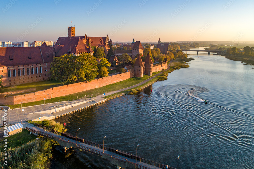 Obraz premium Medieval Malbork (Marienburg) Castle in Poland, main fortress of the Teutonic Knights at the Nogat river, bridges and a water scooter. Aerial view in sunset light.