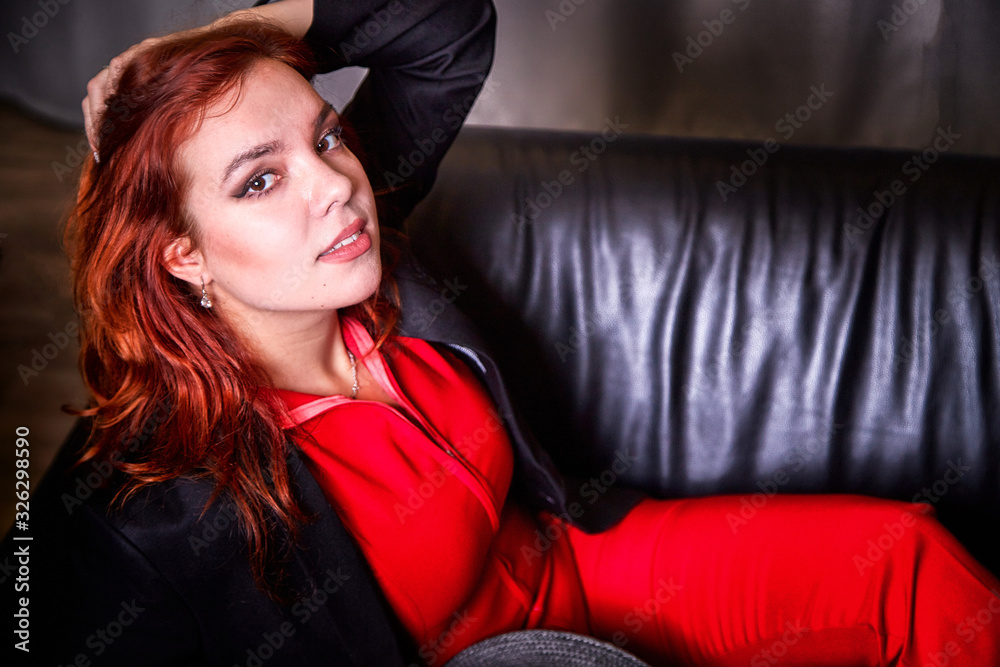 Naklejka premium Beautiful young woman with redhair, black hat and red dress on black sofa in the room and nice light behind her