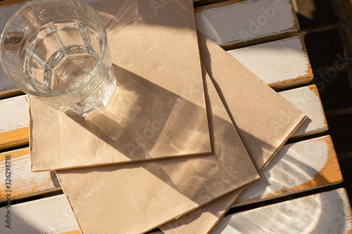 A recycled paper napkins and glass of water are on the cafe's light wooden table in the morning sun.