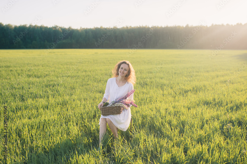 Beautiful curly woman in a white dress holds a basket of flowers in her hands. Against the background of a field with green grass Beautiful curly woman in a white dress holds a basket of flowers in her hands. Against the background of a field with green grass