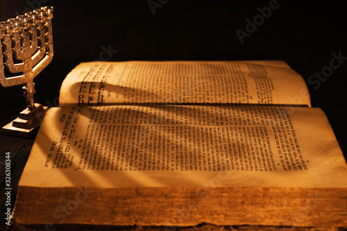 Hebrew Bible and silver menorah in light of burning candle on dark background. Shadow from menorah on open pages of Jewish prayer book in the darkness. Torah reading. Close-up photo