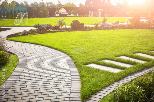 Landscaping of the garden. A tile path between green grass and a lawn with flowers in the sun. Soccer field in the background with copy space.