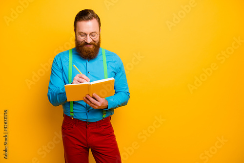 Photo of attractive guy holding writing notes in personal planner have creative idea for new project wear bright blue shirt green suspenders red pants isolated yellow color background
