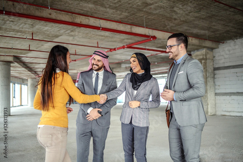 Cheerful attractive arab female investor shaking hands with real estate agent and smiling. Men looking at them and smiling, too. It is time to finally sell old building and transform it to be modern.