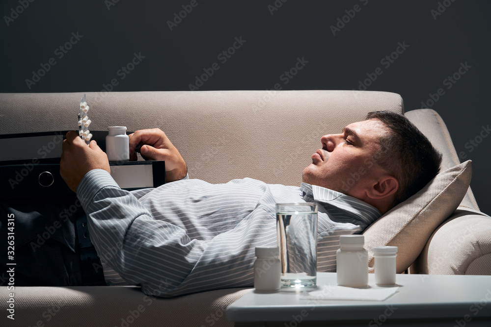 Overworked businessman is lying on the sofa and resting. Pills and glass of water near him. Overworked businessman is lying on the sofa and resting. Pills and glass of water near him.