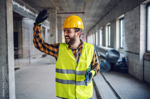 Fotografía Smiling positive construction worker in work wear standing in building and waving to his colleague