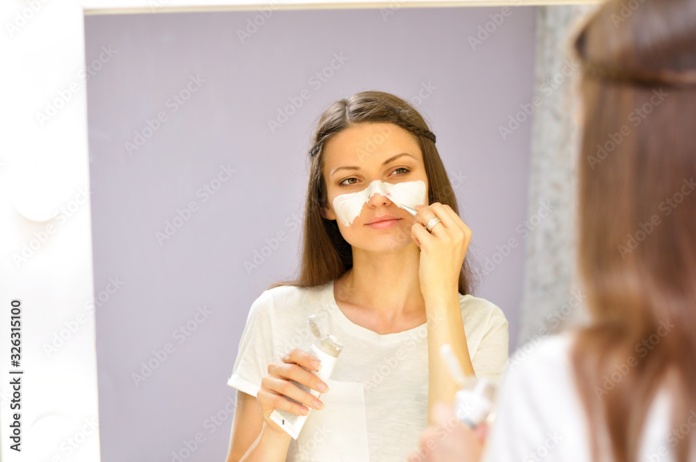 A young beautiful woman puts on a white mask on her cleansed face while standing at home in front of the dressing room mirror in a white T shirt. A young beautiful woman puts on a white mask on her cleansed face while standing at home in front of the dressing room mirror in a white T shirt.