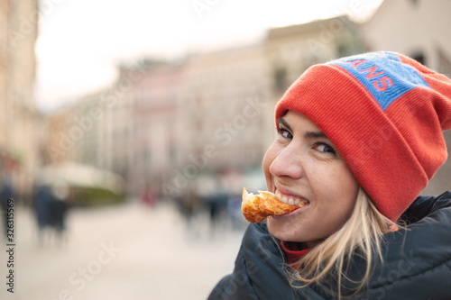 Blonde young woman with naturally beautiful sun kissed face biting a white bread crust and laugh on blurred city background  sunny cold weather