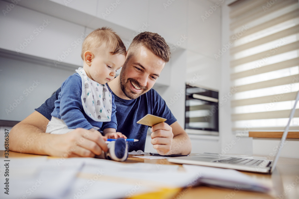 Young attractive bearded father sitting at dinning table with his beloved adorable son and paying bills online. His boy holding pen and trying to help him. Young attractive bearded father sitting at dinning table with his beloved adorable son and paying bills online. His boy holding pen and trying to help him.
