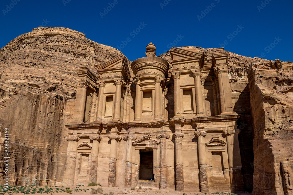 Sunny cloudless day rocky foreground view of the stunning Ad-Deir in the ancient city of Petra, Jordan. Ad-Deir or The Monastery. Petra complex and tourist attraction, Hashemite Kingdom of Jordan