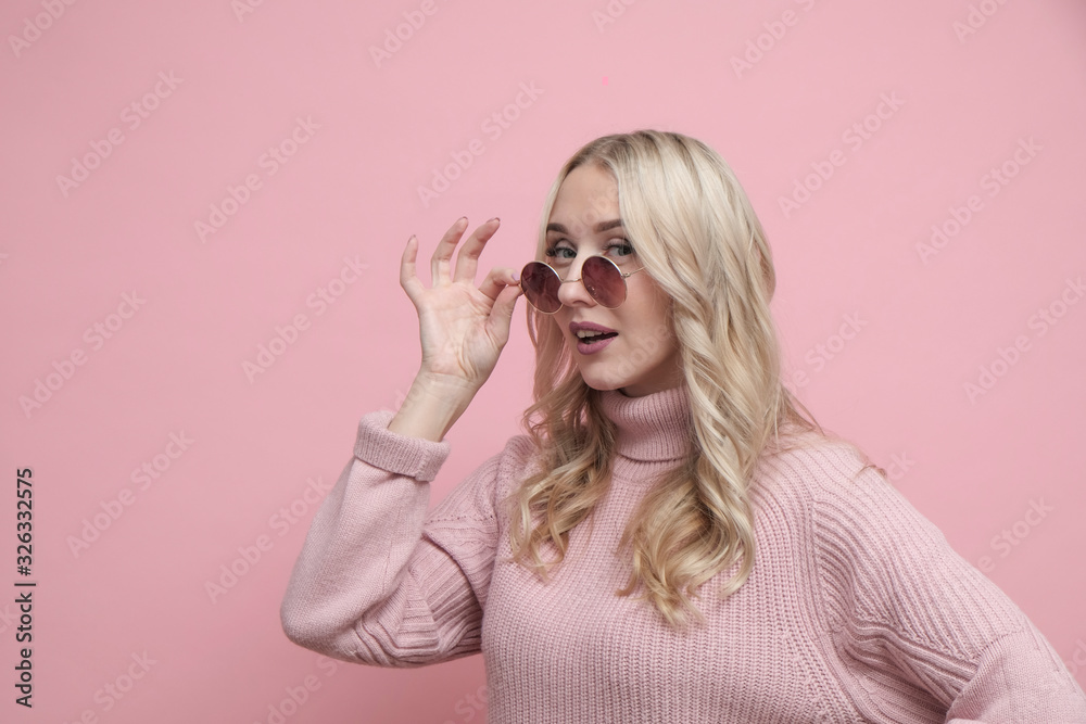 Happy coquettish woman face with blond hair in glasses and wool sweater on pastel pink background. Close up