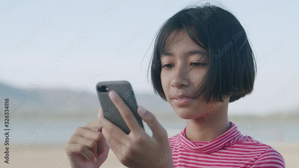 Medium close up of Asian girl using smartphone video call or chat on the beach at sunset, Happy Asia child smile on the face Amid nature. Lifestyle and relaxing concept Medium close up of Asian girl using smartphone video call or chat on the beach at sunset, Happy Asia child smile on the face Amid nature. Lifestyle and relaxing concept
