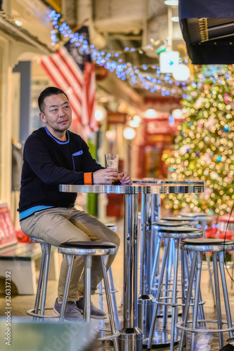 A young Asian man sits at a table in a cafe with a beer glass, with Christmas decorations in the background