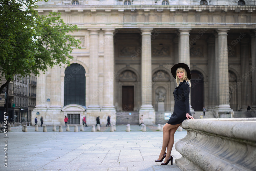 Beautiful long-legged blonde in a short black dress is standing near the fountain in front of Saint-Sulpice church in Paris