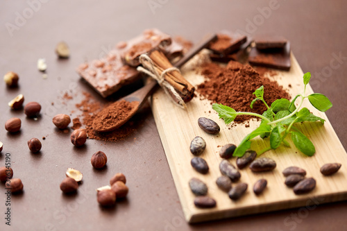 sweets, confectionery and culinary concept - chocolate with hazelnuts, cocoa beans, powder and cinnamon on wooden board with spoon on brown background