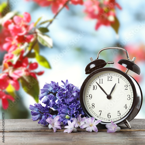 Black alarm clock and flowers on wooden table against blurred background. Spring time