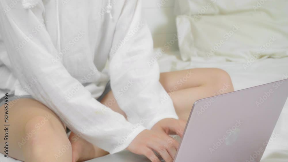 Woman working with her laptop while sitting on a snow-white bed, top view